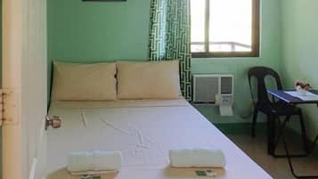 A single bed with two pillows, a bedside table with a chair, a window with patterned curtains, and a wall-mounted air conditioner.