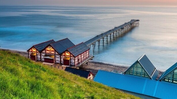 A coastal scene with a pier extending into the sea, a grassy area with a building with multiple rooms, and a modern structure with a sloped roof. - Restored Circus Wagon (Saltburn-by-the-Sea)