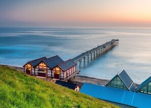 A coastal scene with a pier extending into the sea, a grassy area with a building with multiple rooms, and a modern structure with a sloped roof. - Restored Circus Wagon (Saltburn-by-the-Sea)