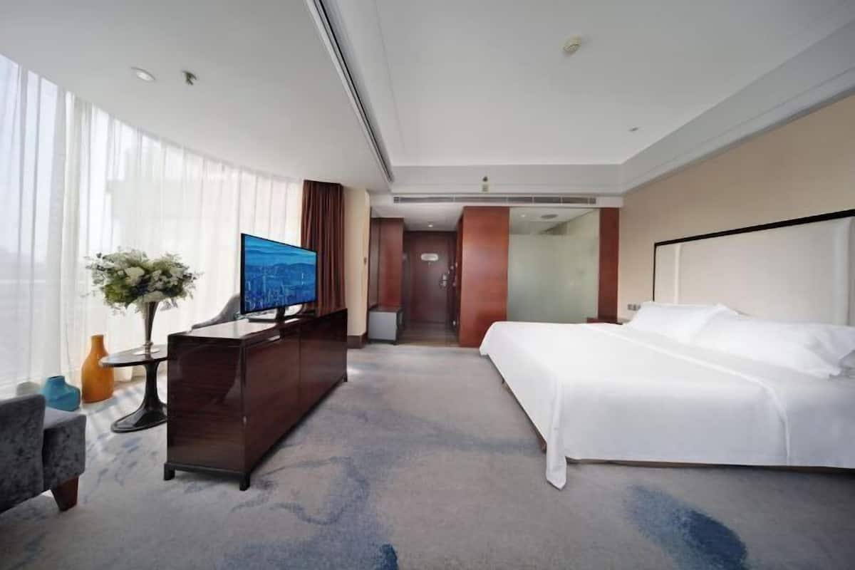 Ein Hotelzimmer mit Bett, Schreibtisch mit Fernseher, Blumenvase und Blick auf die Stadt durch das Fenster.