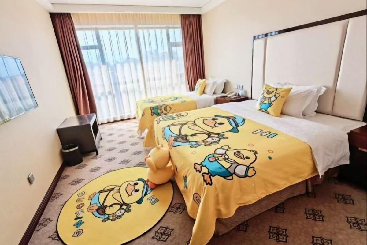 Een hotelkamer met twee bedden, elk met een gele sprei waarop tekenfilmfiguren staan afgebeeld, een groot raam met gordijnen en een vloerbedekking.