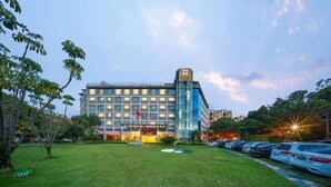 A modern hotel with a glass facade, a parking lot, and a grassy area in front. - Vanilla Garden Hotel (Xiamen)