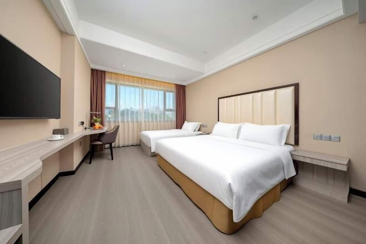 Ein Hotelzimmer mit zwei Betten, einem Schreibtisch, einem Stuhl, einem Flachbildfernseher und einem großen Fenster mit Vorhängen.