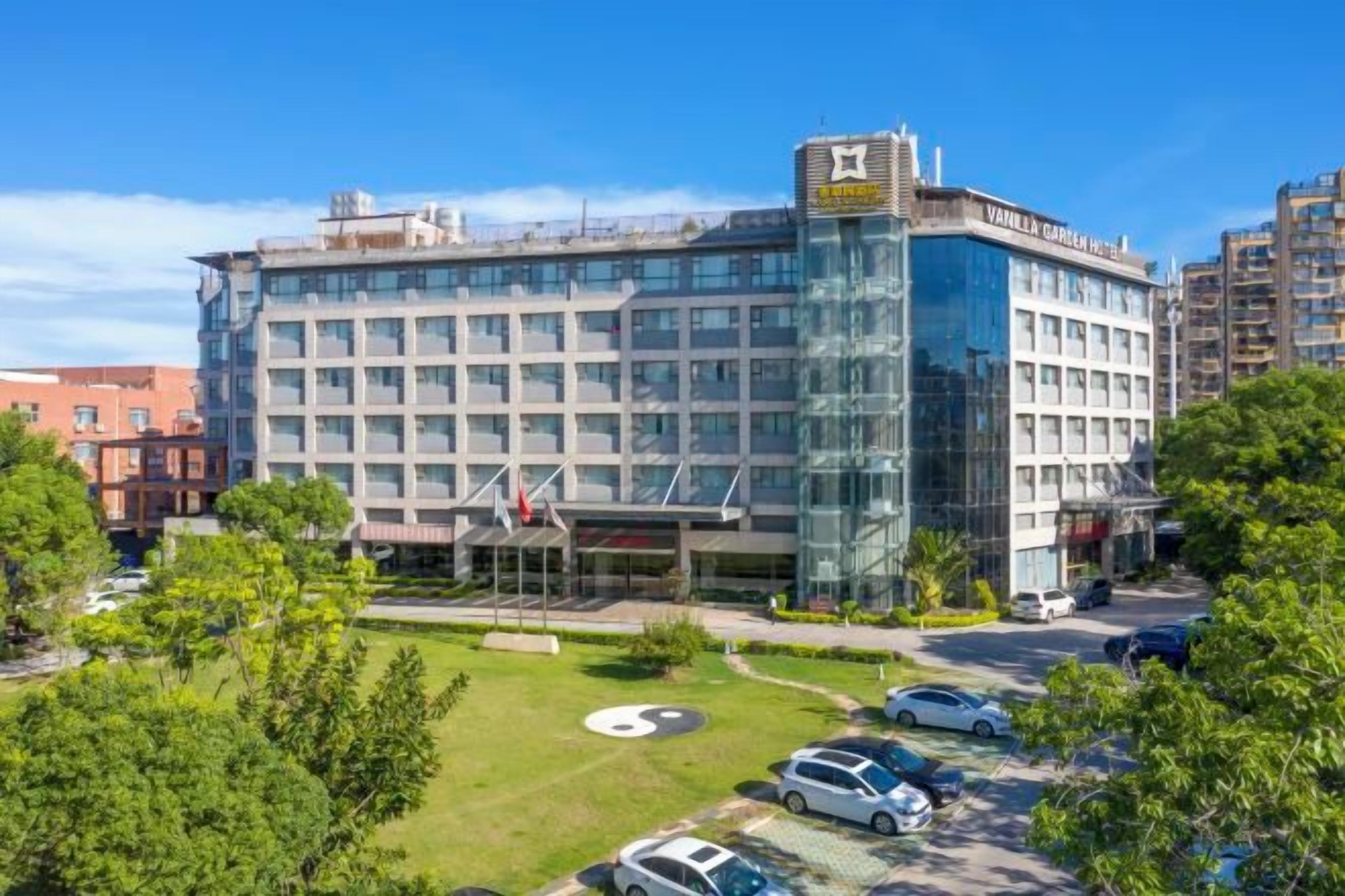 A modern hotel with a glass facade, multiple floors, and a parking area with cars.