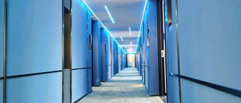 A long, narrow hallway with blue walls and a patterned floor, leading to a bright area at the end.
