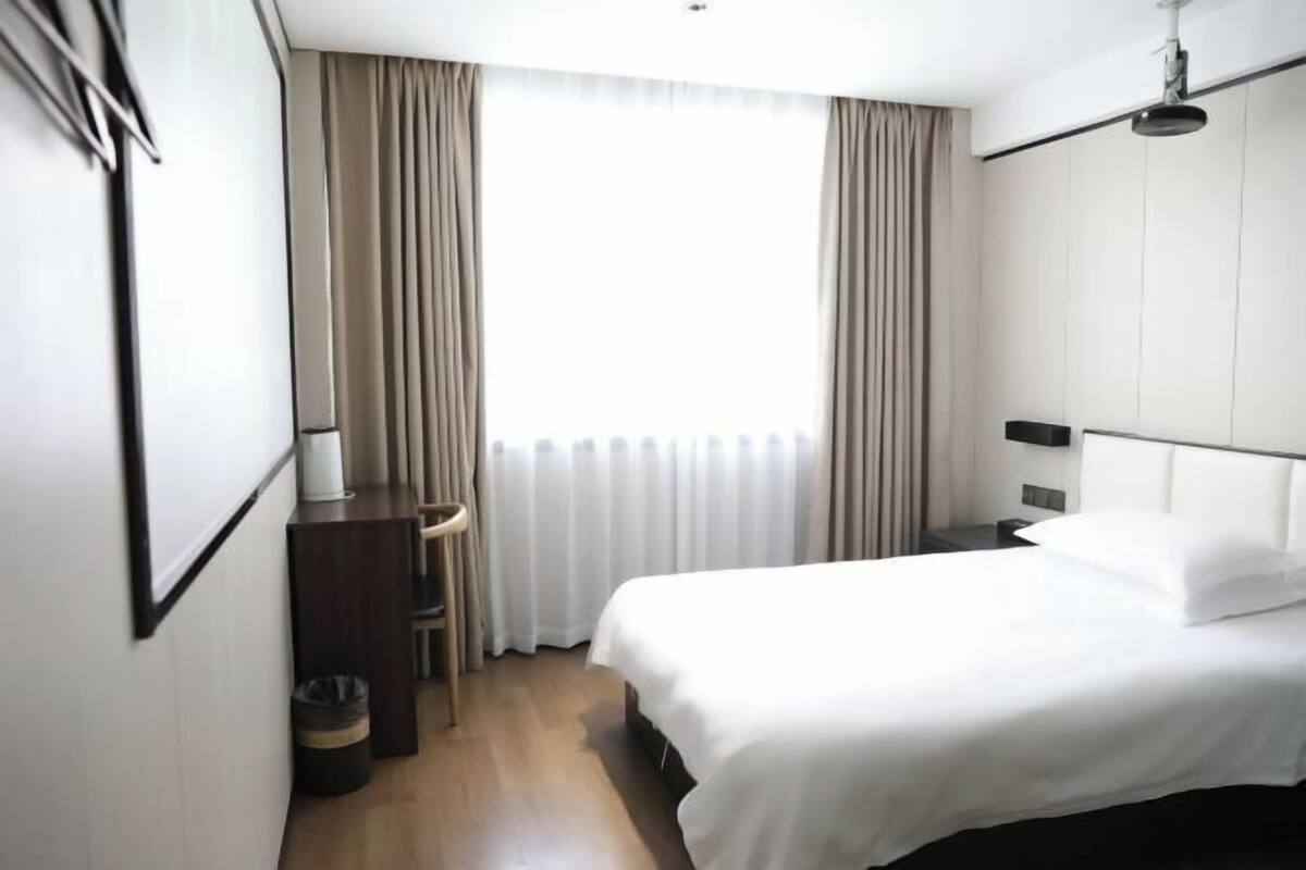 Una camera d'albergo con un letto, una scrivania, una sedia, un cestino e una finestra con tende.