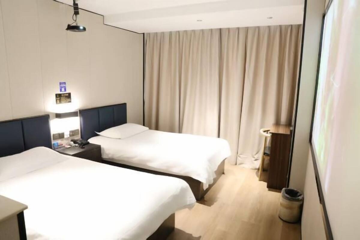 Una camera d'albergo con due letti, un comodino, una lampada, una tenda e un cestino.