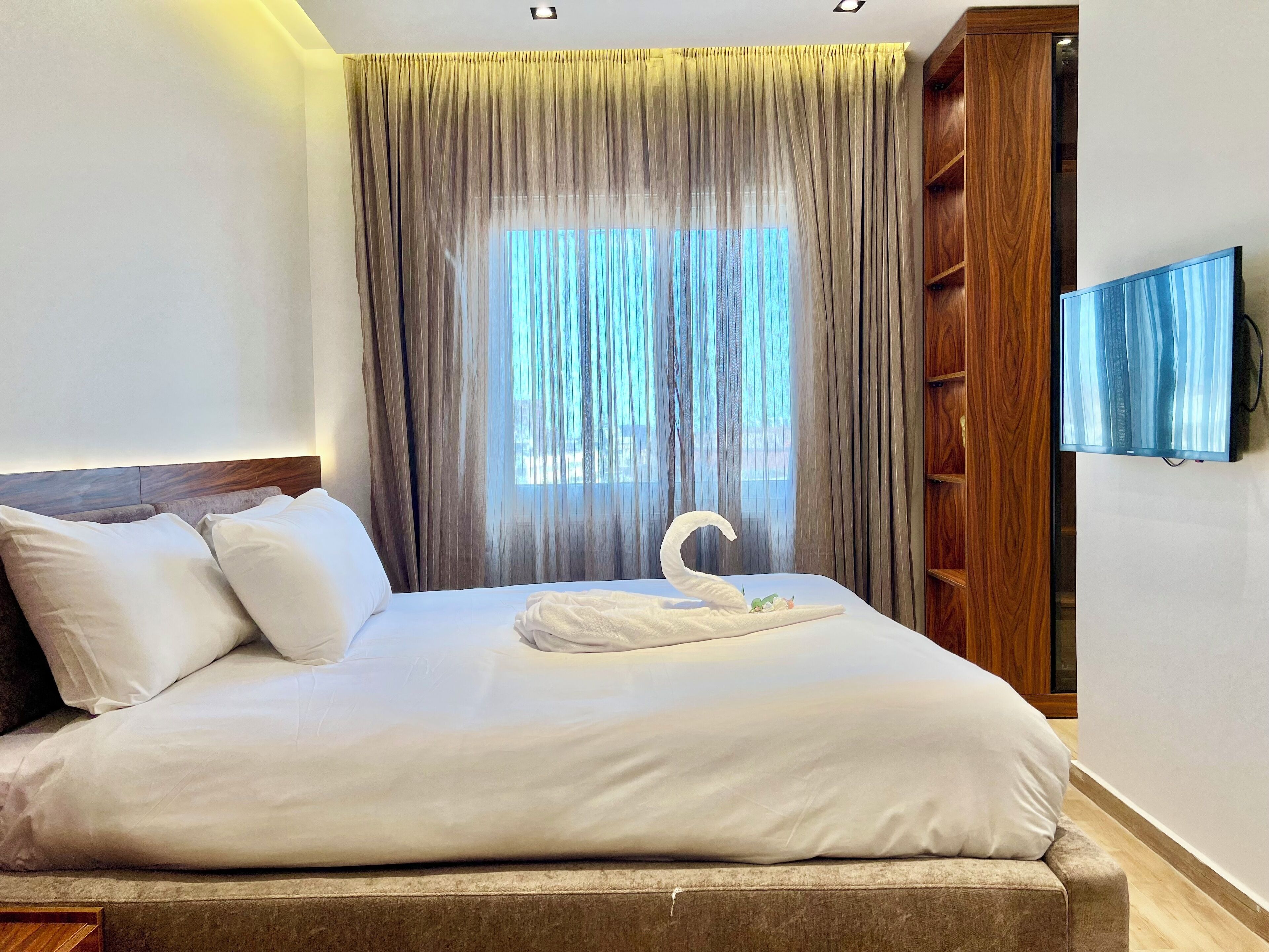 A neatly made bed with white linens, a wooden headboard, and a swan-shaped pillow. There is a large window with sheer curtains, a wooden bookshelf, and a flat-screen TV mounted on the wall.