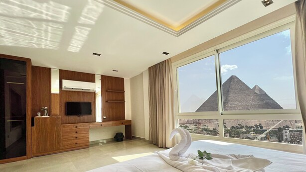 A hotel room with a bed, a TV, a wardrobe, and a large window with a view of pyramids. - Gaia Pyramids View INN (Giza)