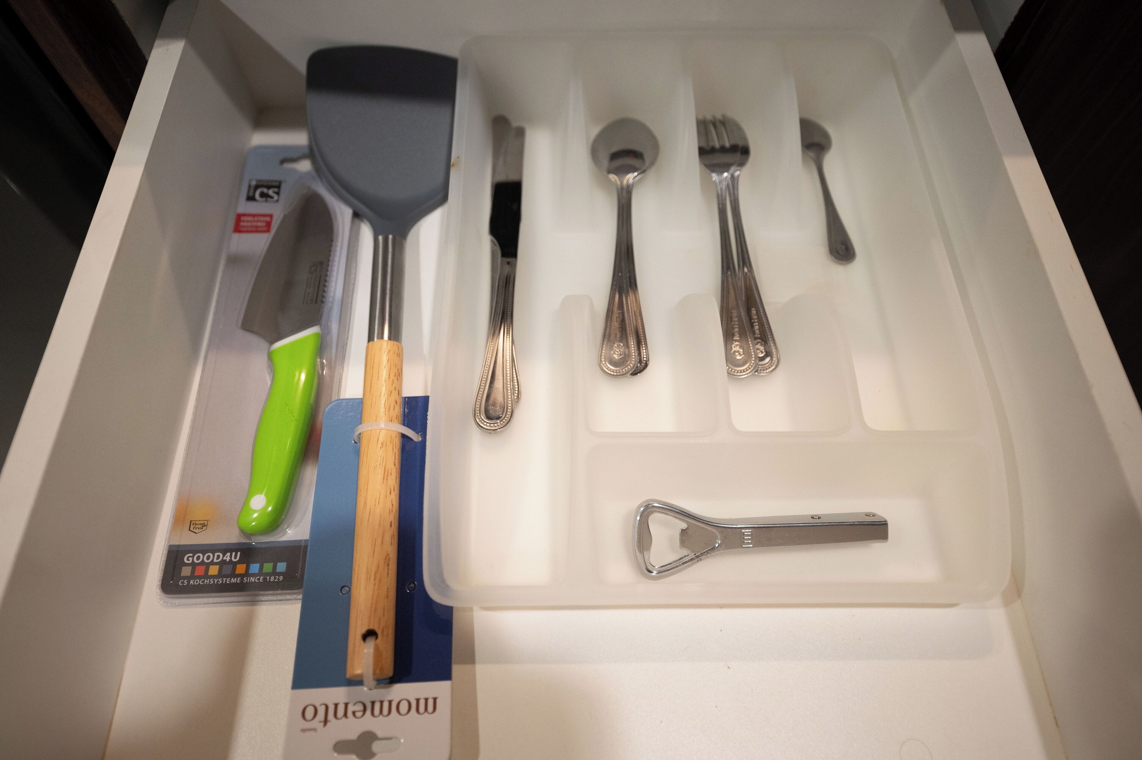 Un tiroir contenant une spatule à manche vert, une spatule à manche en bois, une cuillère, une fourchette et un décapsuleur.