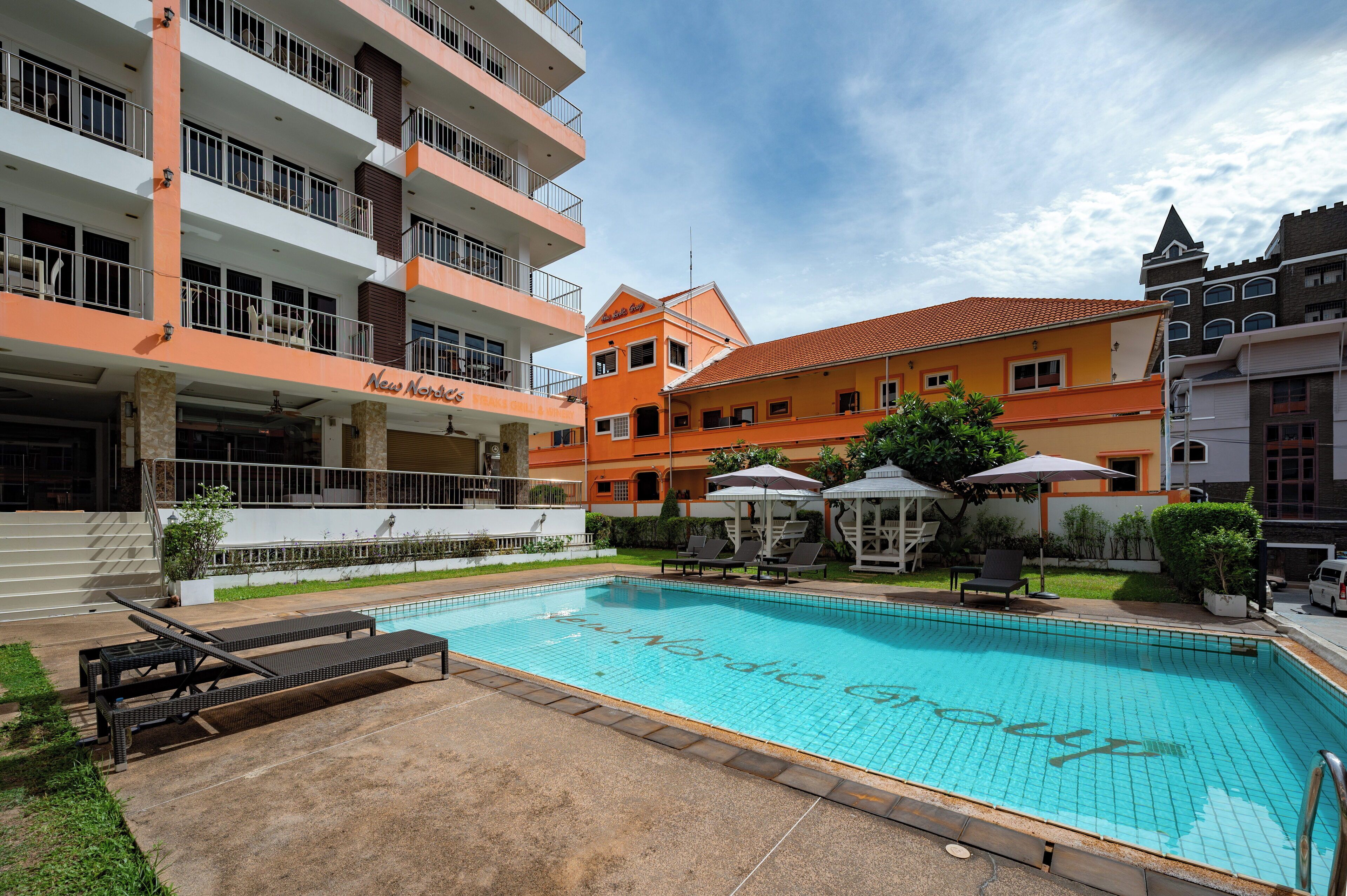 A swimming pool with lounge chairs and umbrellas, surrounded by multi-story buildings with balconies and outdoor seating areas.