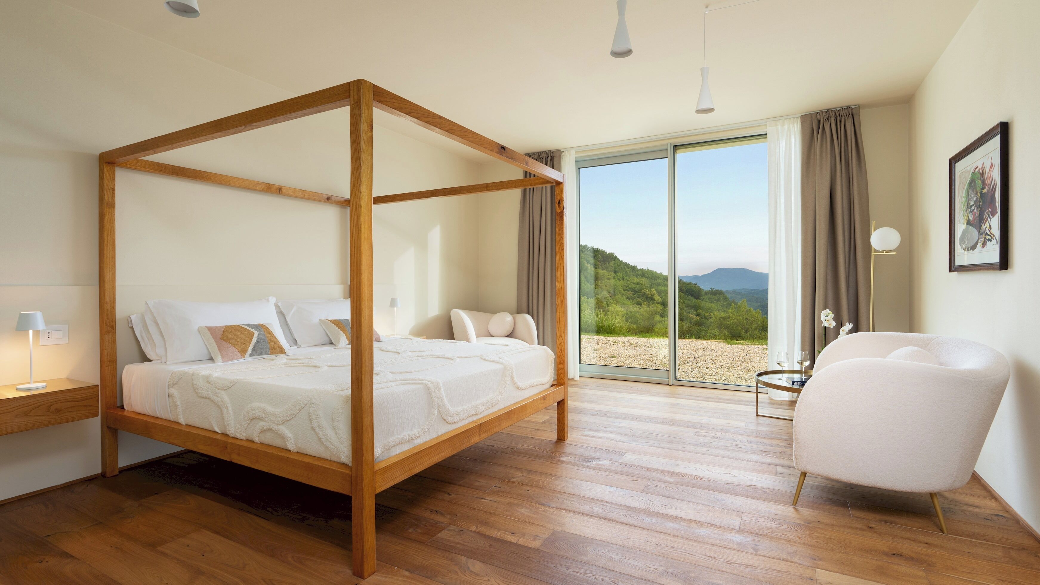 A bedroom with a four-poster bed, a sitting area with a chair, a small table, and a view of a natural landscape through a large window.