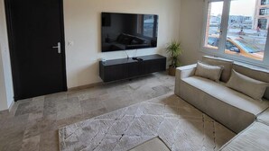 Un soggiorno moderno con una televisione a schermo piatto, un mobile TV scuro, un divano beige e un'ampia finestra con vista sull'esterno.