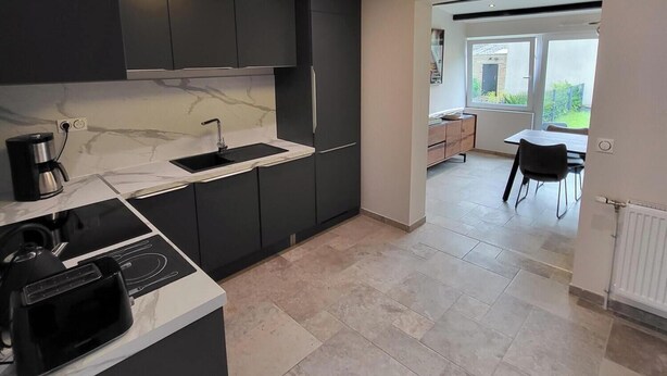 Una cucina moderna con mobili scuri, un piano di lavoro in marmo e un piano cottura integrato. Si intravede una zona pranzo con tavolo e sedie attraverso un'apertura.