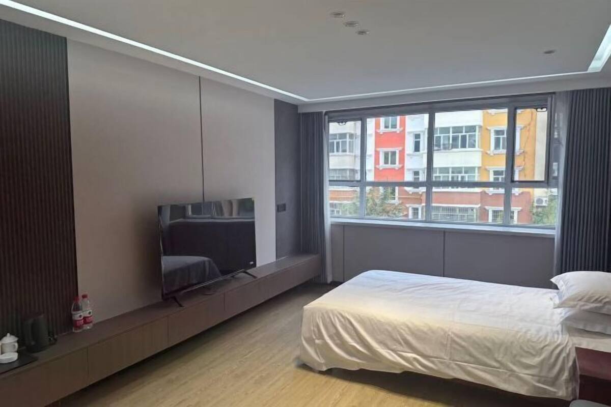 A modern hotel room with a bed, a TV, a built-in bench, and a large window with a view of buildings.