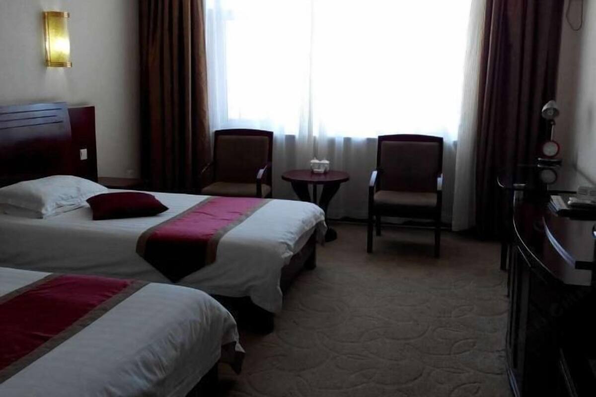 A hotel room with two beds, a small table, two chairs, a desk, and a window with curtains.