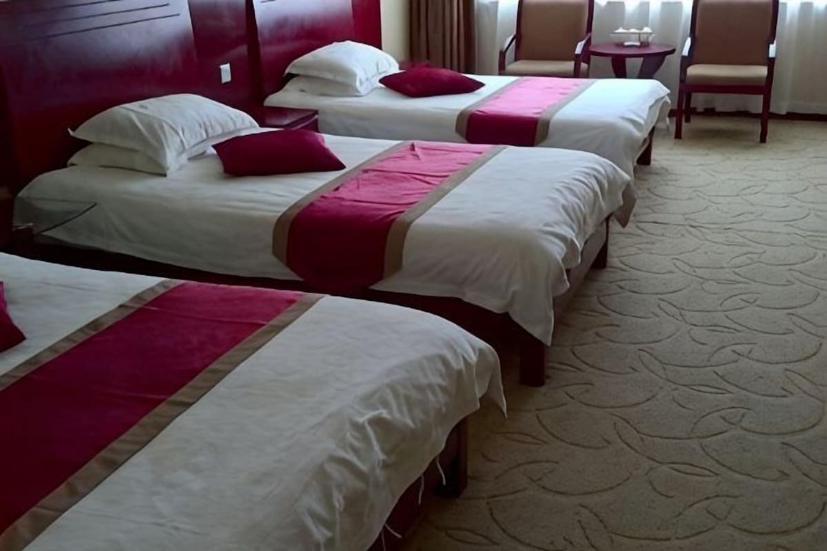 Quarto de hotel com três camas, cada uma com roupa de cama branca e detalhes vermelhos, piso coberto por carpete e uma mesa pequena com uma cadeira.