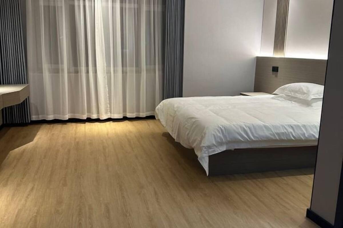 A hotel room with a bed, a bedside table, a curtain, and a wooden floor.
