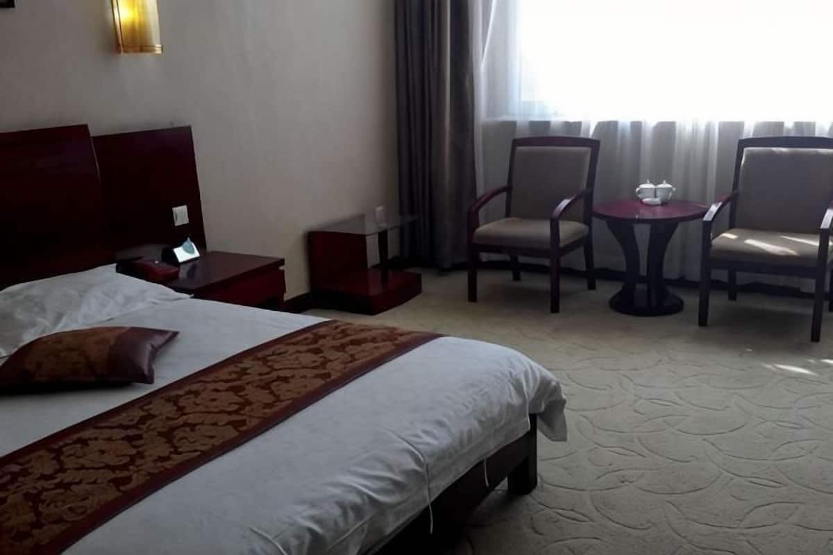 A hotel room with a large bed, a bedside table, a small table with chairs, and a window with curtains.