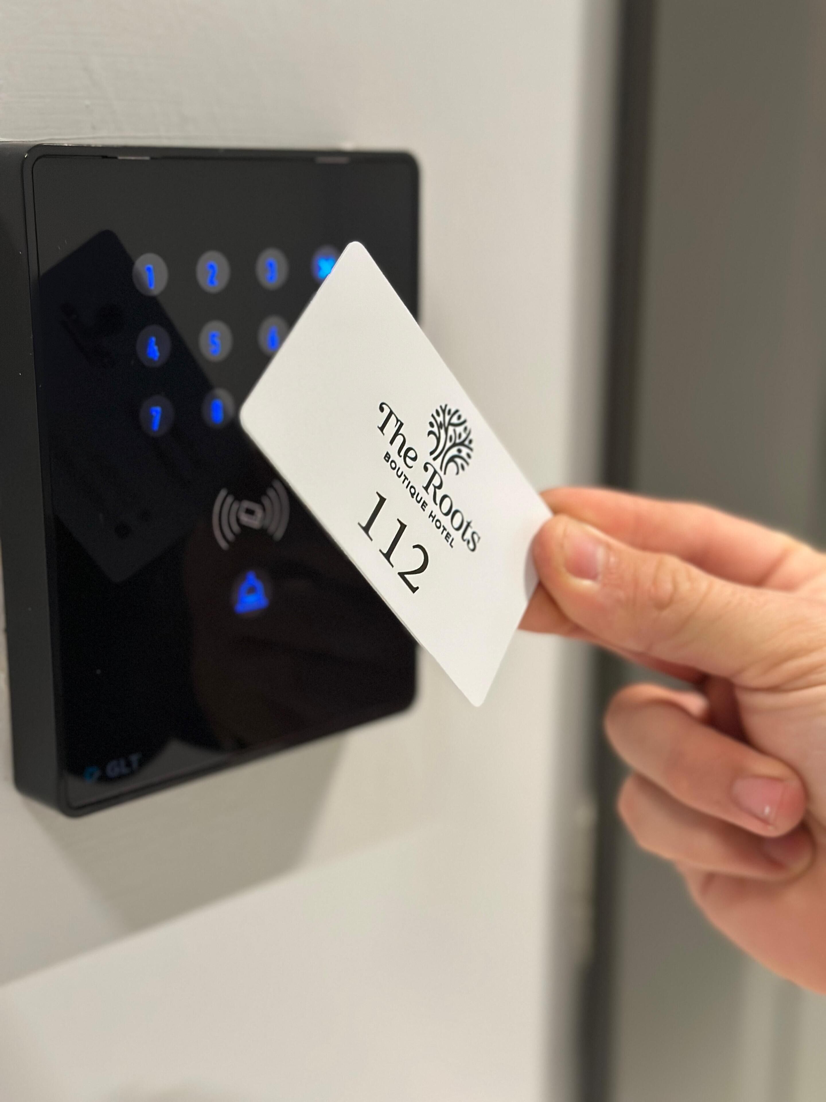 A hand holding a card with "The Roots Boutique Hotel 112" printed on it, in front of a digital keypad.