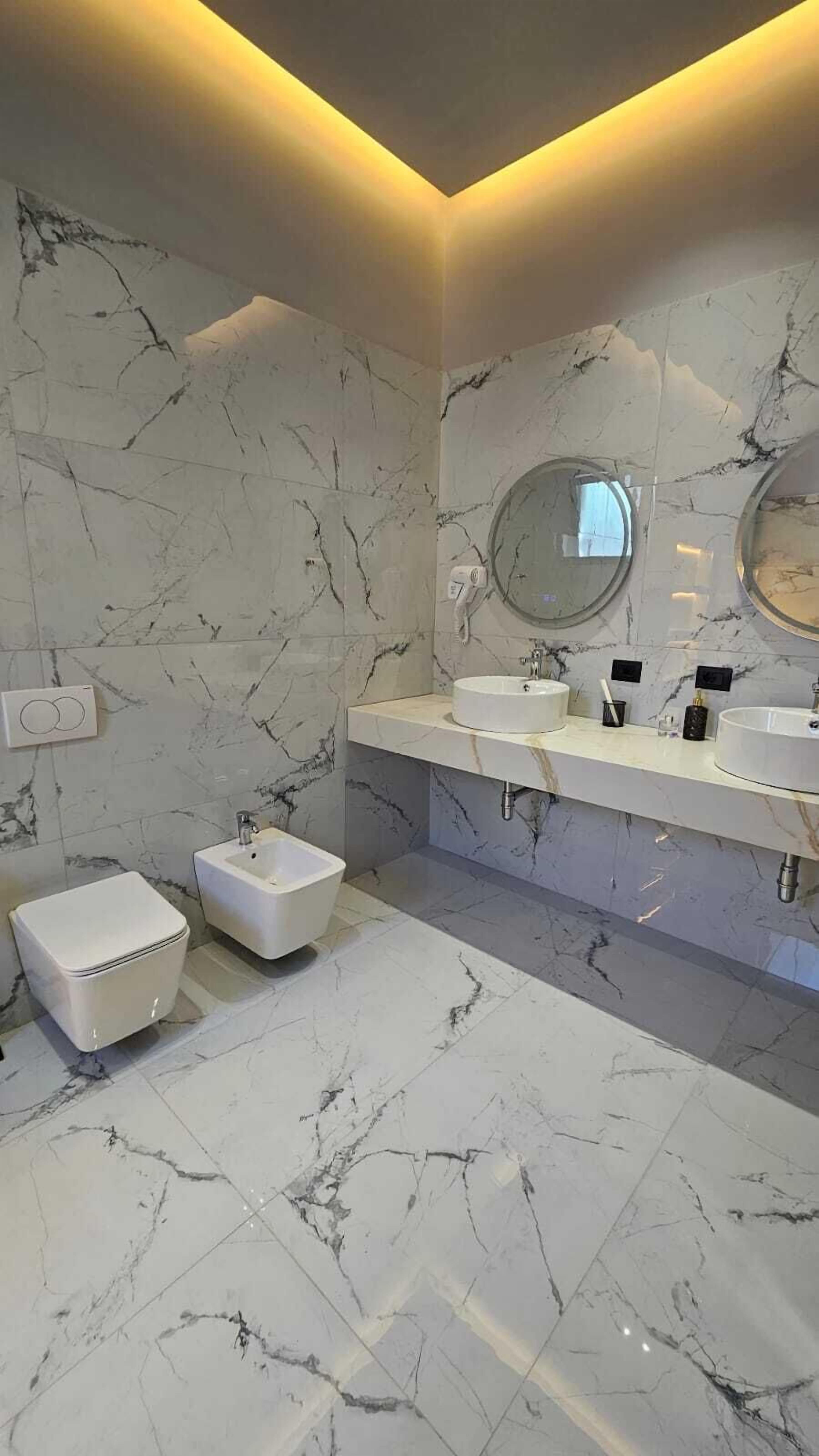 A modern bathroom with marble walls and floors, a round mirror, two sinks, and a small white pedestal sink.