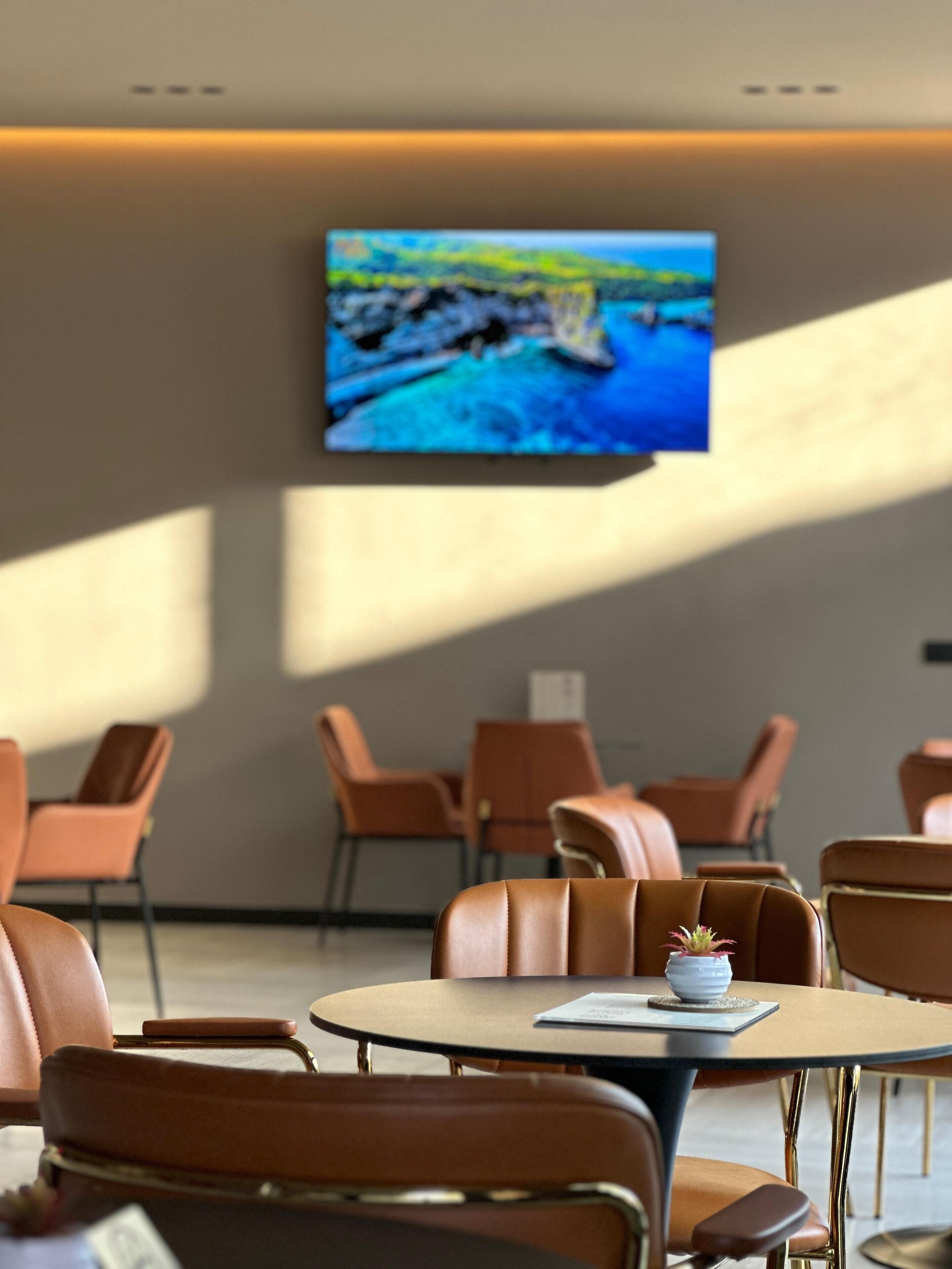 A modern dining area with leather chairs and a round table, a flat-screen TV mounted on the wall, and a view of a rocky coastline.