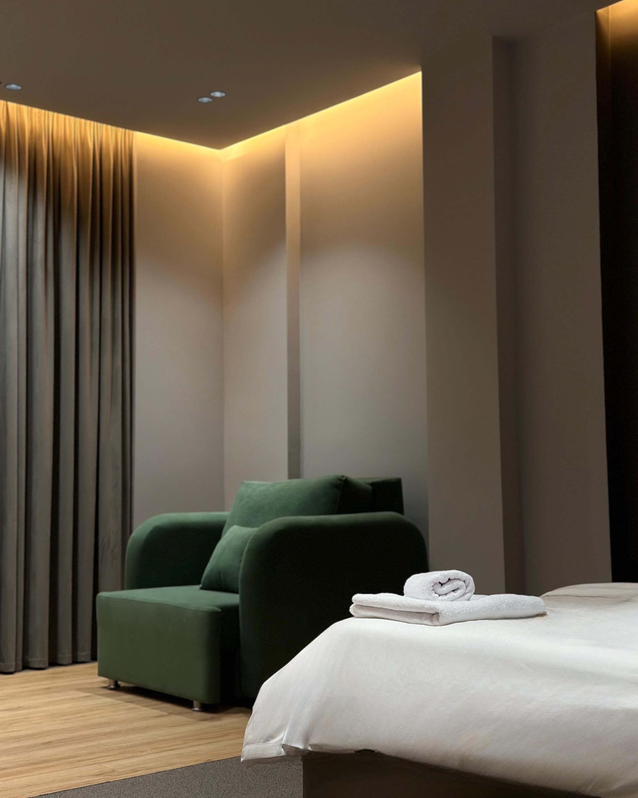 Una habitación de hotel moderna con un sillón verde, una cama bien hecha y un ventanal con cortinas.