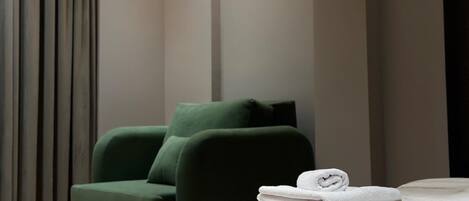 Una habitación de hotel moderna con un sillón verde, una cama bien hecha y un ventanal con cortinas.