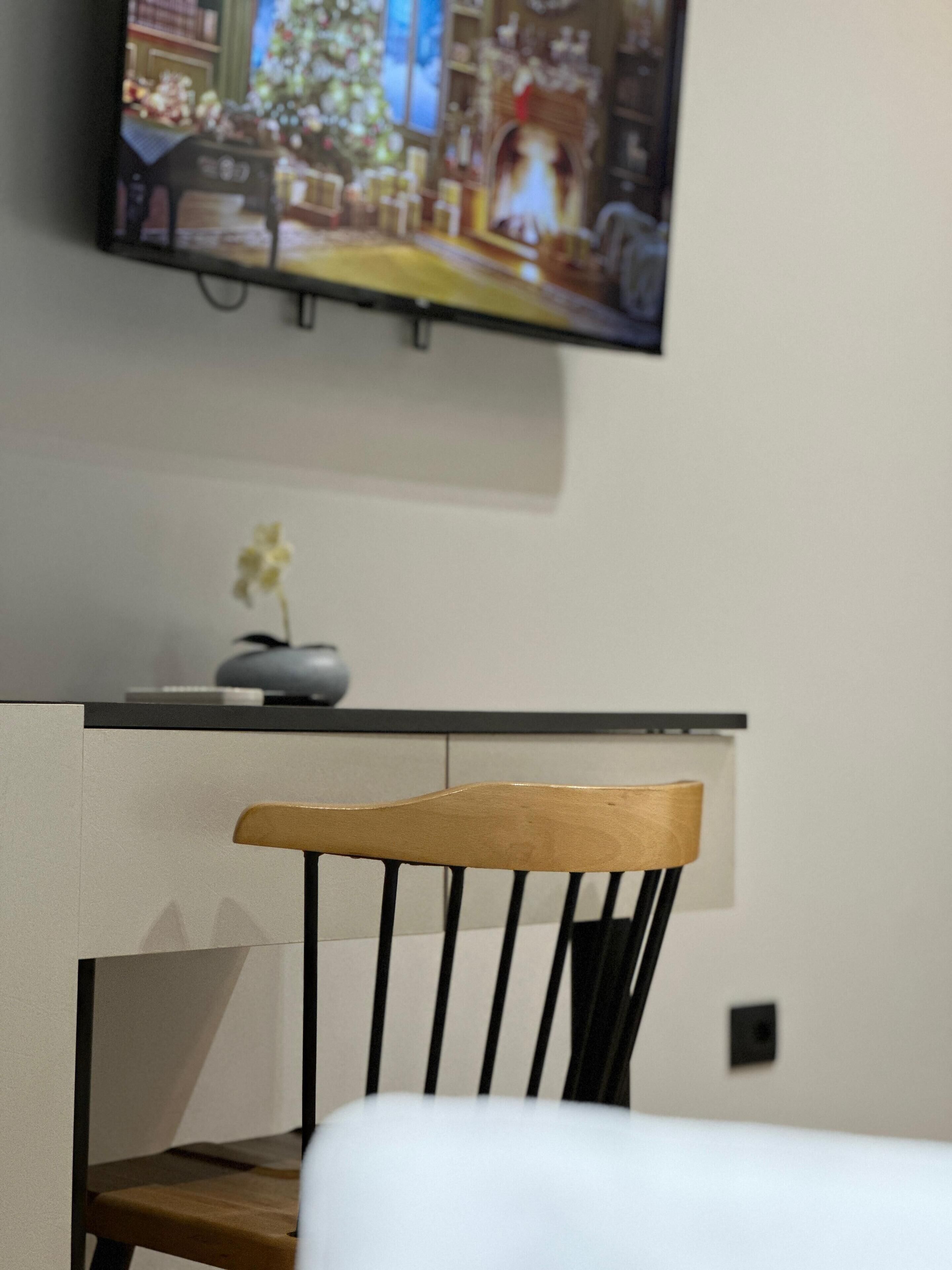 A chair with a wooden backrest and black legs, a small vase with a single flower, and a television mounted on the wall displaying a scene with a Christmas tree.