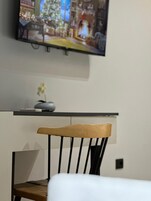 A chair with a wooden backrest and black legs, a small vase with a single flower, and a television mounted on the wall displaying a scene with a Christmas tree.