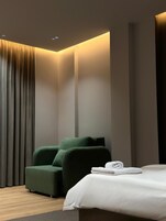 A modern hotel room with a bed, a green armchair, and a large window with vertical blinds.