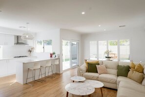 Eine moderne Küche mit weißen Schränken, einer zentralen Kochinsel mit Barhocker und einem Essbereich mit einer Couch und zwei runden Tischen.