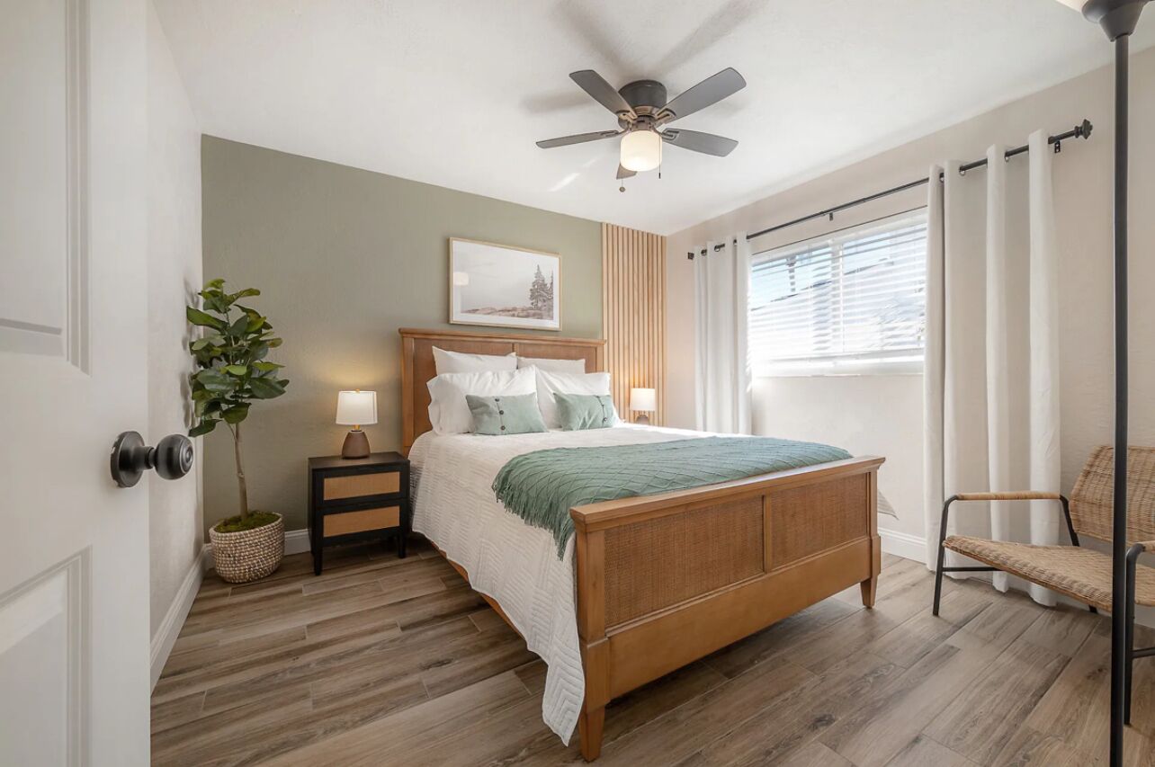 Une chambre à coucher avec un lit en bois, une table de chevet avec une lampe, un ventilateur de plafond, une fenêtre avec des rideaux et une chaise.