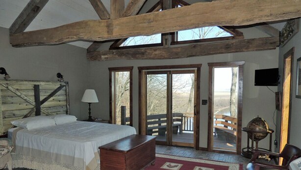 Un dormitorio con techo de madera, una cama, un aparador, una lámpara y vistas al exterior a través de una puerta.