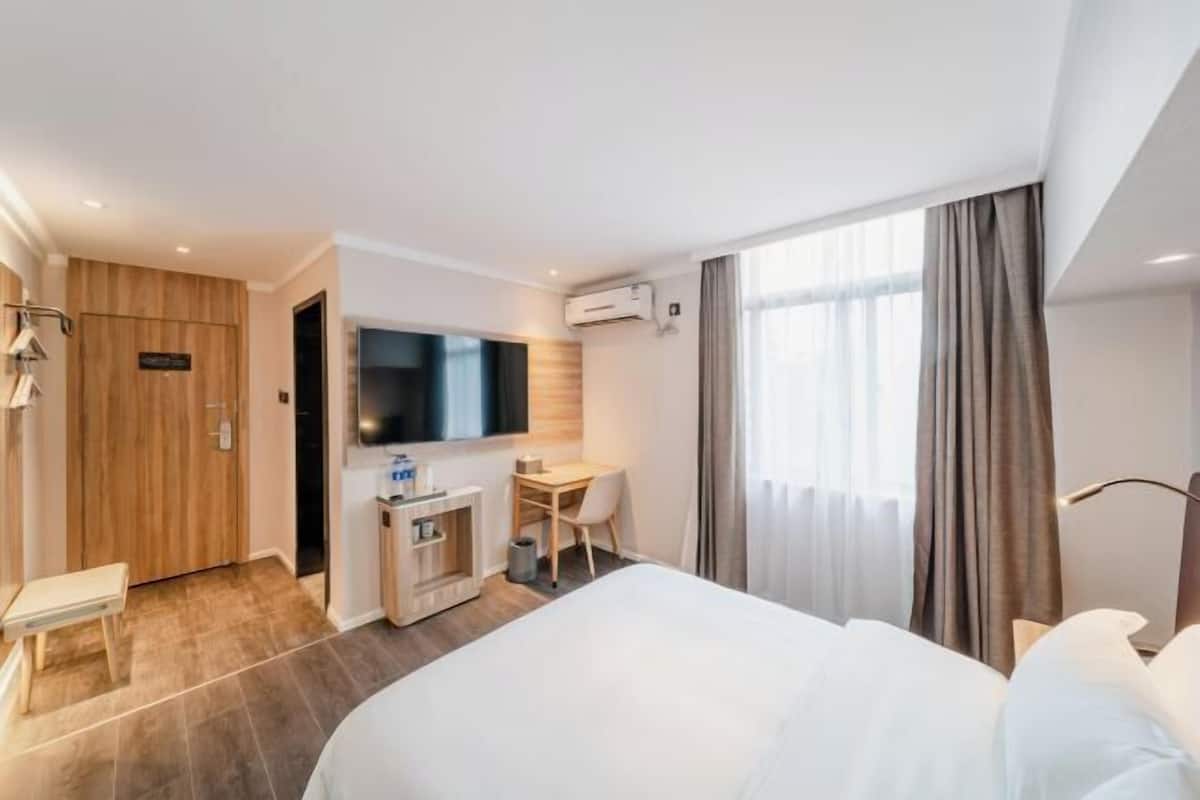 A hotel room with a bed, a desk, a chair, a TV, and an air conditioning unit.