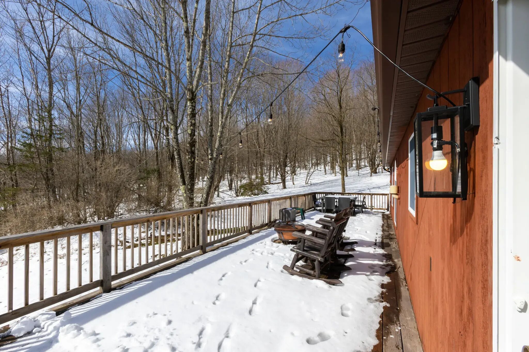 A wooden deck with outdoor seating and a railing, surrounded by snow-covered ground and trees.