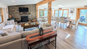 Un salon spacieux avec une cheminée en pierre, un grand téléviseur et un coin repas comprenant des chaises et une table en bois.
