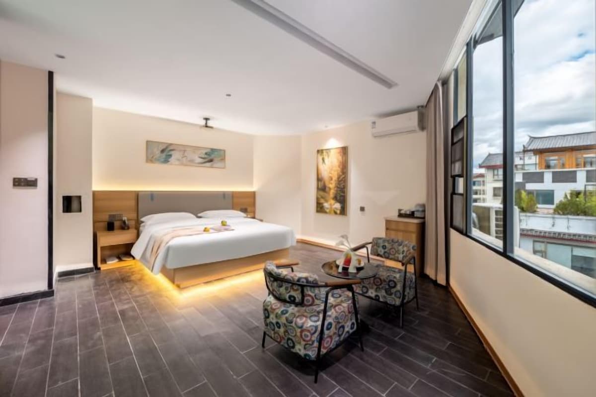 Une chambre d’hôtel moderne, dotée d’un grand lit, de deux fauteuils, d’une petite table et d’une grande fenêtre donnant sur la ville.