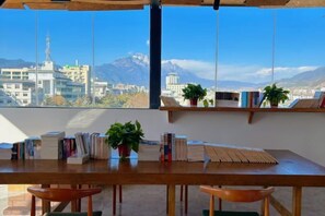 A wooden table with books and plants, a view of a cityscape and mountains through large windows.