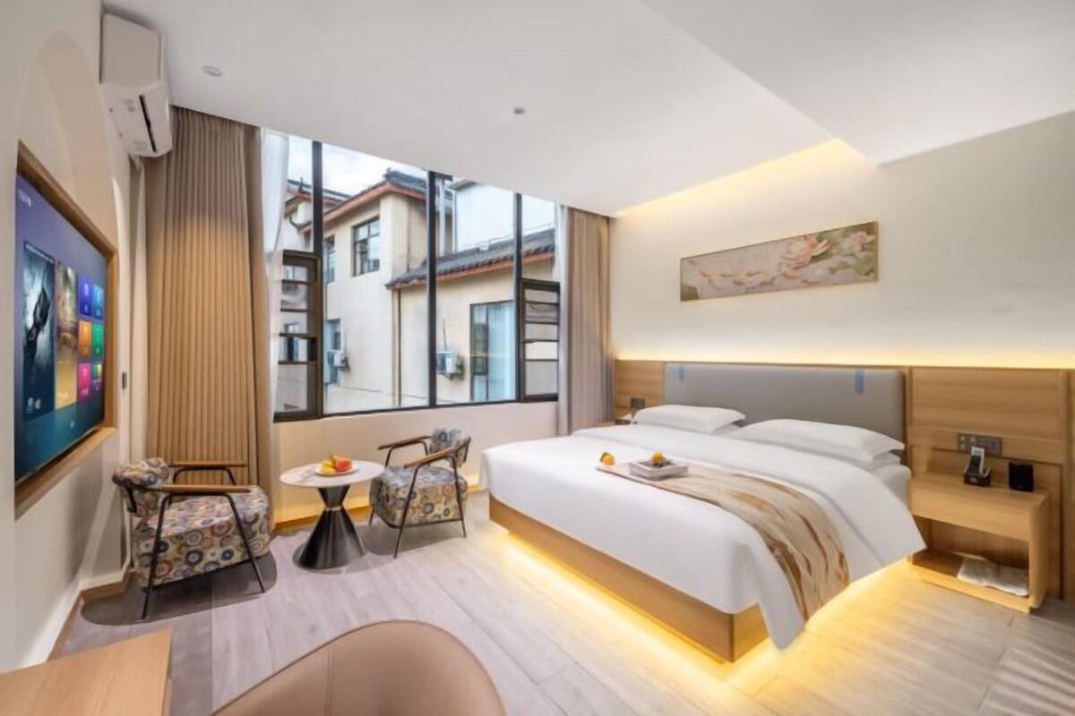 Une chambre d’hôtel moderne dotée d’un grand lit, d’un bureau, de deux chaises, d’une petite table et d’une grande fenêtre donnant sur la ville.