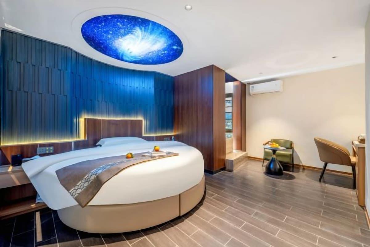 Une chambre moderne avec un lit aux lignes courbes, un plafond décoré sur le thème de la galaxie et un petit coin salon avec une table.
