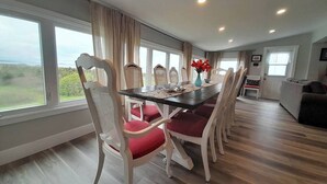 Een eetruimte met een lange tafel en zes stoelen, grote ramen met gordijnen en uitzicht op de omgeving.