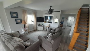 Een ruime woonkamer met een grijze zithoek, een plafondventilator, een trap en een eethoek op de achtergrond.