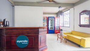 A reception area with a wooden desk labeled "ayenda HOTELES," a yellow sofa, and a window with blinds. - Hotel Puerto Vigia Inn (Cartagena)