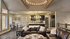 A spacious living room with a large brown leather sofa, a round ottoman, and a dining area with a wooden table and benches. A kitchen with dark cabinets and a central island is visible in the background. - Beautiful Mountain Home, Ski In/Ski Out & Golf within Boyne Mountain Resort (Boyne falls)