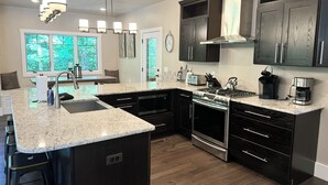A modern kitchen with dark cabinetry, a granite countertop, stainless steel appliances, and a large window providing natural light. - Beautiful Mountain Home, Ski In/Ski Out & Golf within Boyne Mountain Resort (Boyne falls)