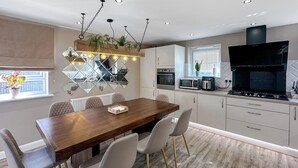 A modern kitchen with a dining area, featuring a wooden table, white chairs, and a built-in oven. - Sheffield Charming Detached 4bd Home (Catcliffe)