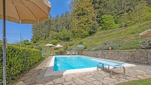 A rectangular swimming pool with stone tiles, surrounded by a stone wall and a grassy area. There are umbrellas and lounge chairs in the background. - Villa in Cortona with 3 bedrooms sleeps 6 (Cortona)