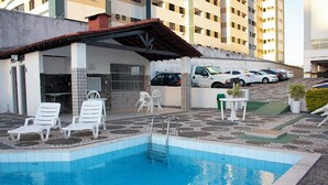 Piscina com espreguiçadeiras brancas, área coberta com teto cerâmico e estacionamento com vários carros.