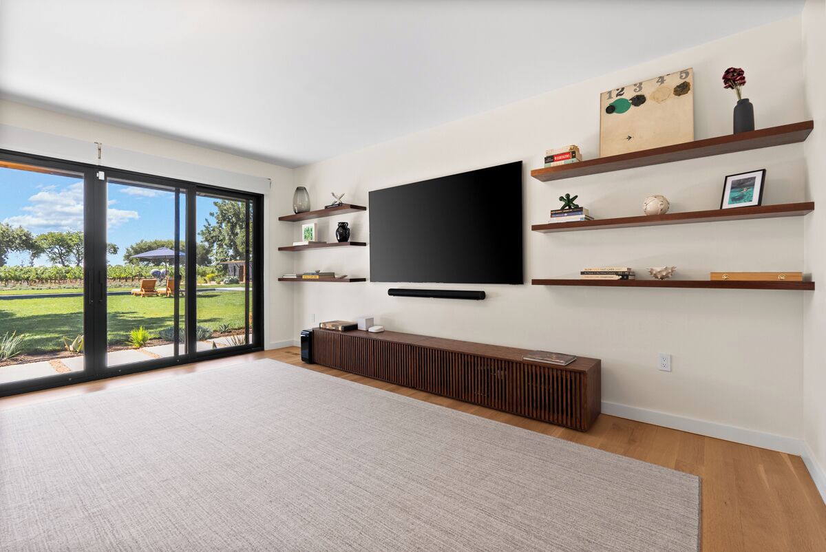 Un soggiorno moderno con una grande TV a schermo piatto fissata al muro, mensole in legno con oggetti decorativi, un mobile console in legno integrato e un'ampia porta scorrevole in vetro che dà su un giardino.